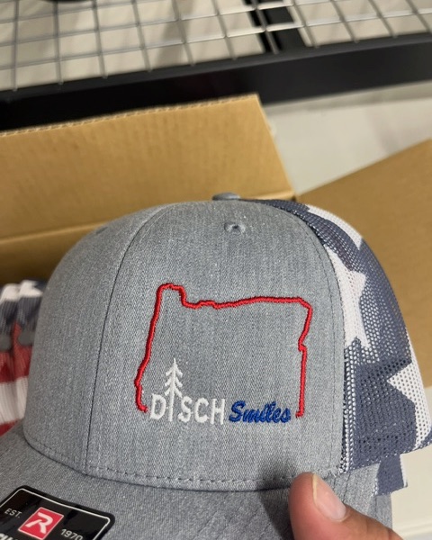 Disch Smiles Oregon outline logo embroidered in red, white, and blue on a heather gray Richardson trucker cap