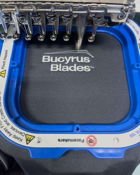 Bucyrus Blades logo stitching in white on a black shirt, held in a blue magnetic embroidery hoop