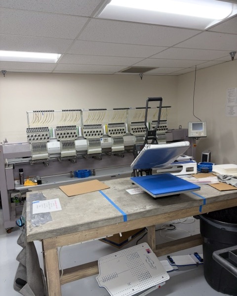 Heat press on a work table in front of a multi-head embroidery machine at Serena Embroidery